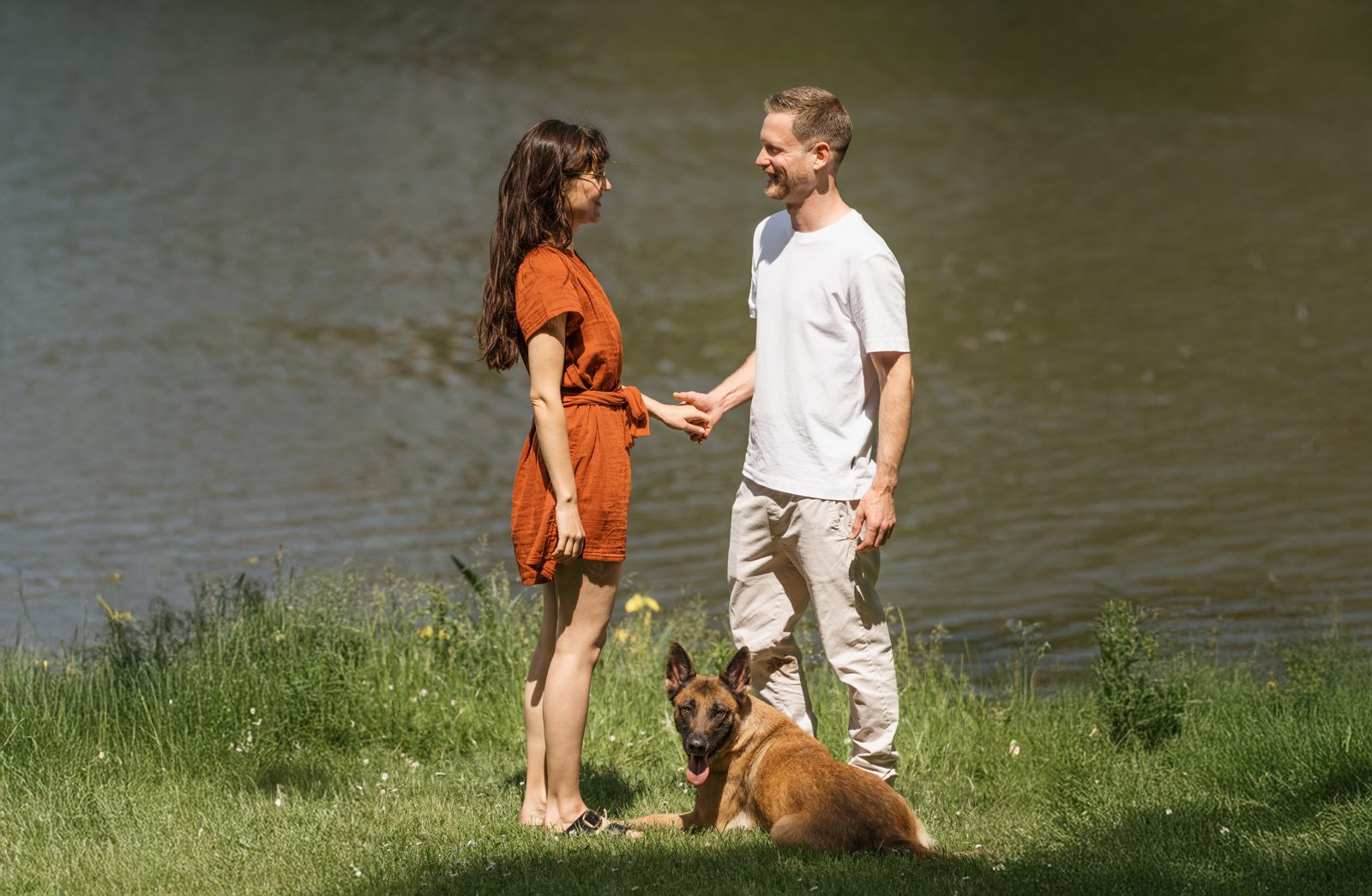 Ronja und Clemens stehen sich am See gegenüber und halten Hände, Mali sitzt zwischen ihnen